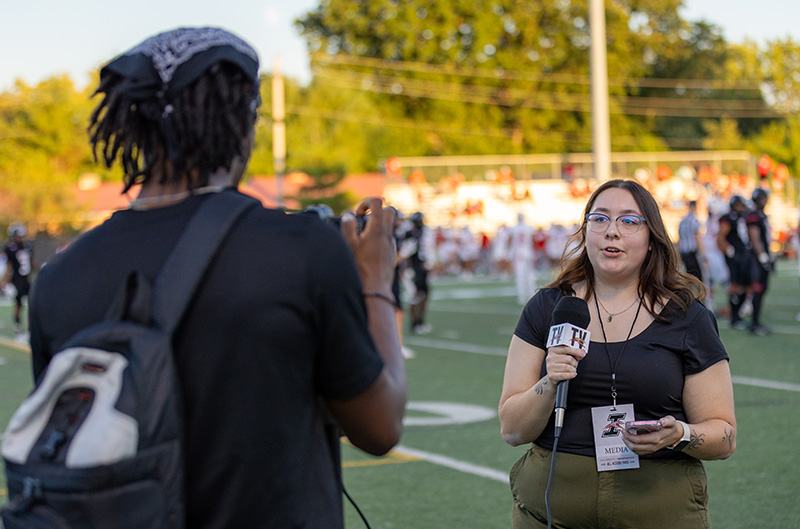at a football game, one student is holding a microphone while another is filming