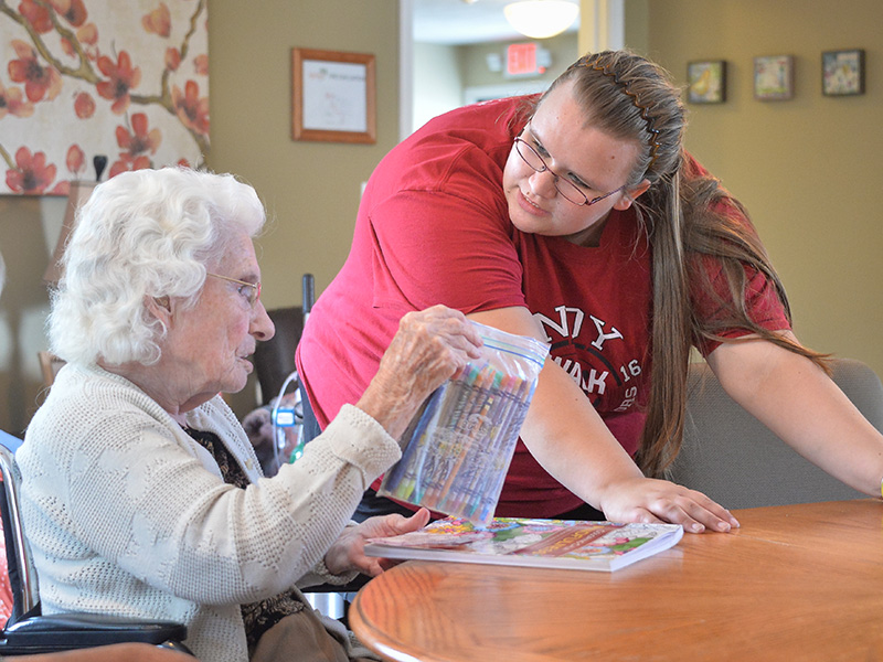 A student converses with an elderly patient as they prepare to color in a coloring book. 