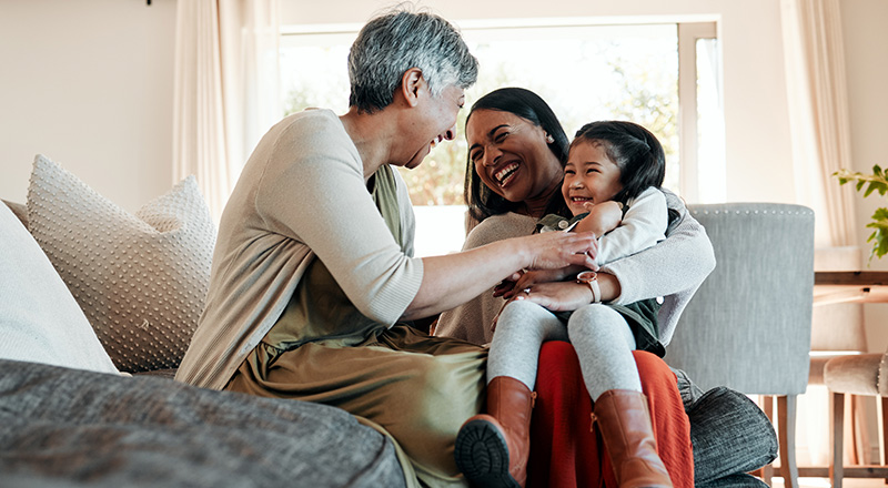 A grandmother, mother, and young daughter are laughing on the sofa