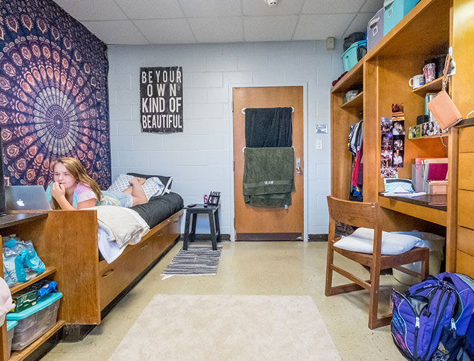 different angle of a girl on her laptop on her bed. on the other side of the room is closet, desk, and dresser.