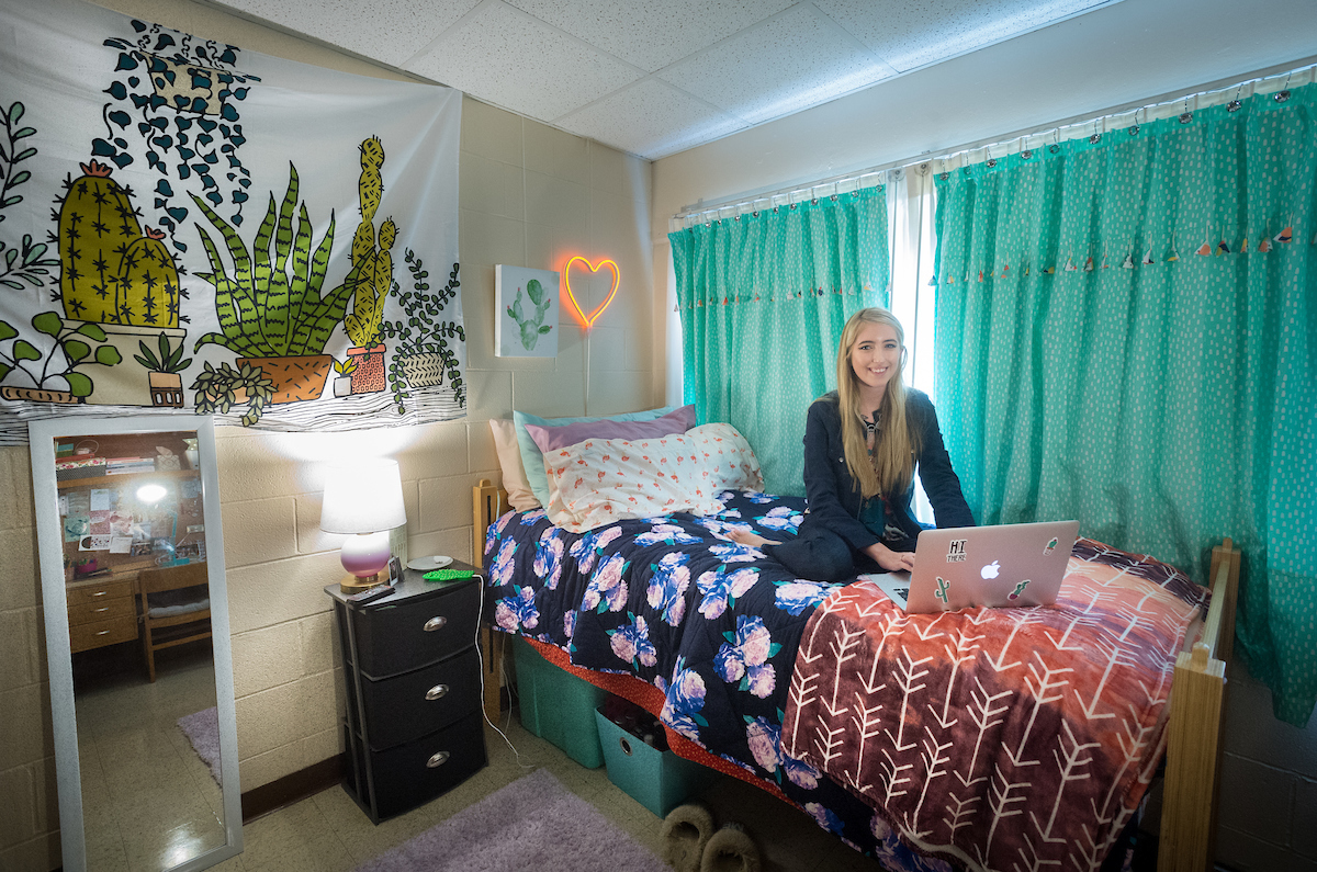 a girl is on her laptop sitting on her bed next to a lamp, night stand, and window with curtains.