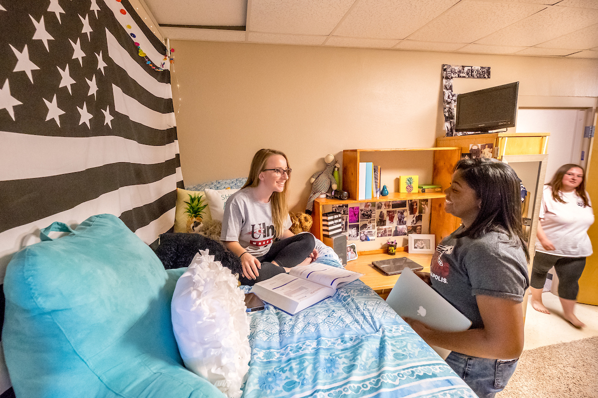 two female students talking. one is sitting on her bed, the other is standing. there is a desk beside her bed decorated with several photos and books.