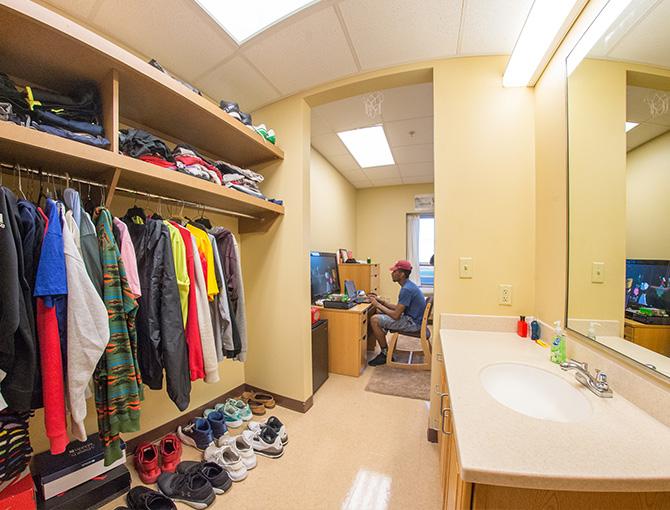 bathroom with sink/mirror on one side and doorless closet on the other side. in the distance is a student on his laptop sitting at his desk. 